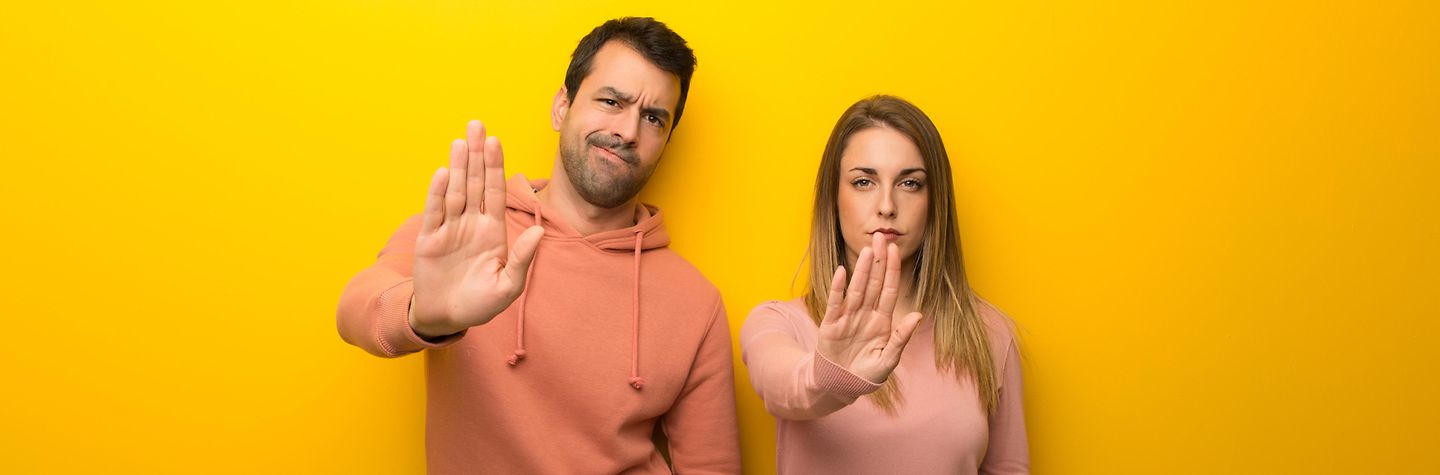 Zwei Personen vor gelbem Hintergrund. Beide zeigen eine Stop-Geste mit der rechten Hand. Ihr Gesichtsausdruck ist abwehrend.